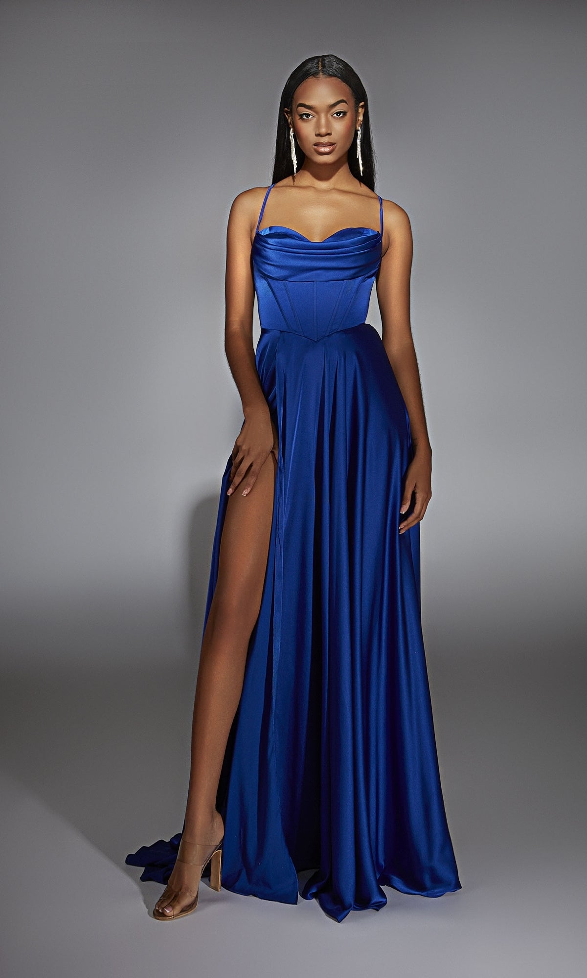 A woman poses in a studio wearing the Alyce 61883 Long Sweetheart A-Line Prom Dress in royal blue, featuring a corset bodice, thigh-high slit, thin straps, heeled sandals, and drop earrings.